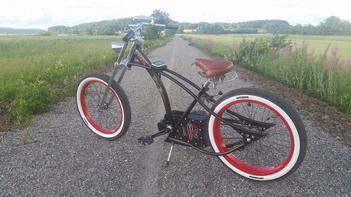 Custombyggd elcykel på en grusväg med breda rödvita däck, brunt sadel och distinkt batteriboxdesign, omgiven av fält och natur.