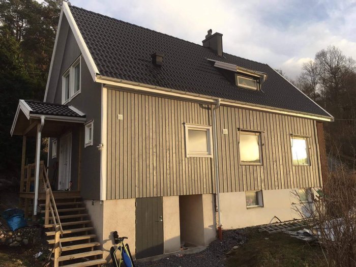 Tvåvåningshus med lutande tak och liggande träpanel, omgiven av natur i skymningen.