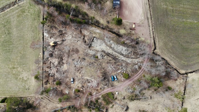 Drönarbild över ett byggprojekt med grävmaskin på en avskogad tomt omgiven av gröna fält.