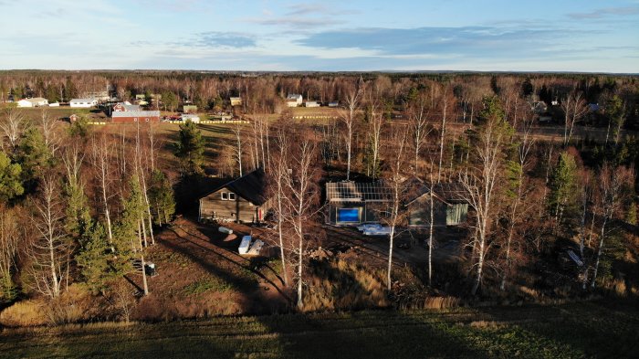 Drönarvy av byggprojekt i skogsmiljö med hus och grundplatta, soligt väder.