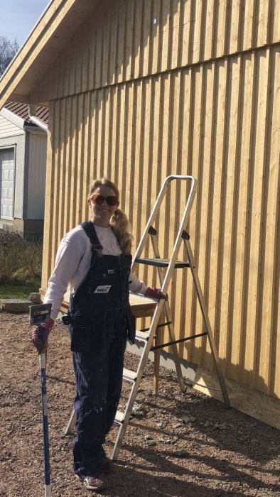 Kvinna i äldre arbetskläder ler framför ett trägästhus under konstruktion med stege vid sidan.