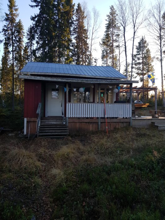 Röd stuga med vit knut och altan som sjunker, omgiven av skog och en svensk flagga.