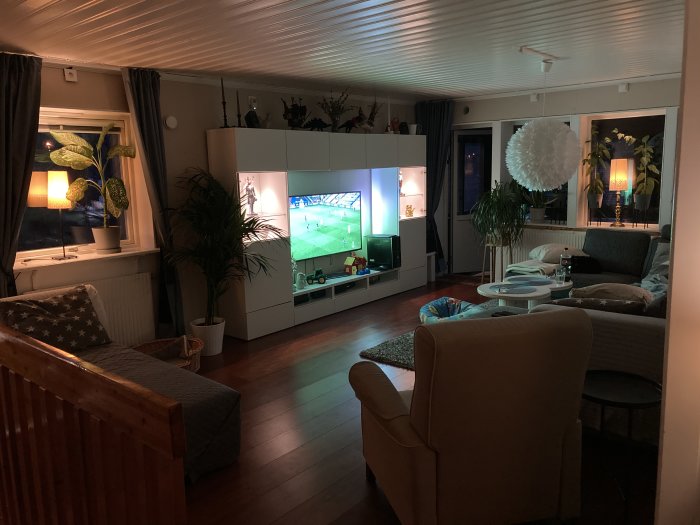 Vardagsrum före renovering med möbler och tv påslagen, indirekt belysning och växter.