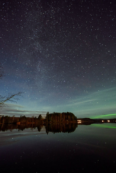 Stjärnhimmel med Norrsken över spegelblank sjö och silhuett av träd och hus i natten.