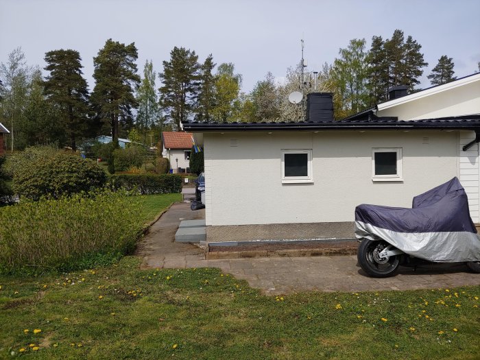 Ett vitt enplansgarage med svart tak och tre små fönster, till höger en motorcykel under presenning.