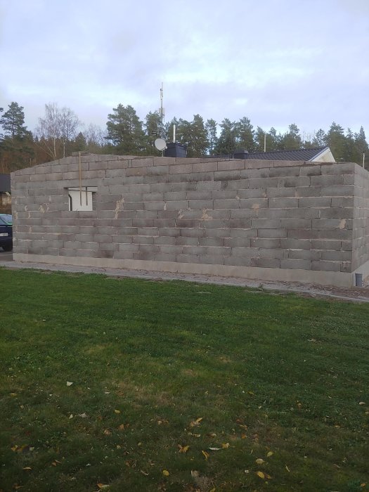 Trehövdad grå lecablocksvägg under uppbyggnad mot befintligt garage, med gräsmatta i förgrunden.