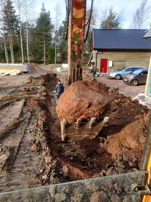 En grävmaskin som lyfter en stor sten ur en rörgrav på en stenig tomt med en person som iakttar.