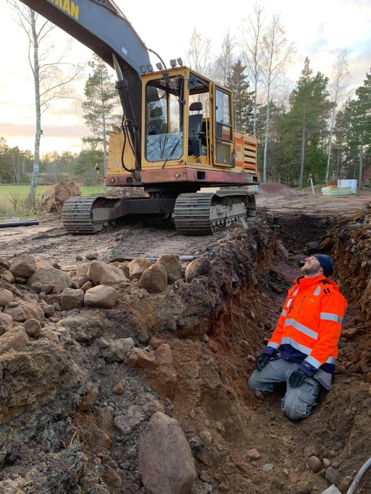 Man i skyddskläder sitter i grävd rörgrav bredvid en grävmaskin på en byggarbetsplats.