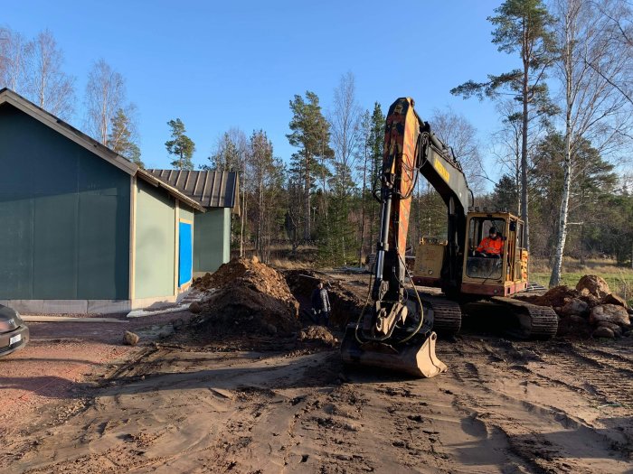 Grävmaskin vid hus och garage arbetar med markarbeten under solig himmel, omgivet av stora stenar och högar av grus.