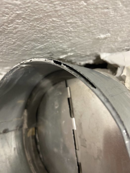 Närbild på en skadad vägg där en böjd ventilationskanal delvis kommer fram genom ett hål.