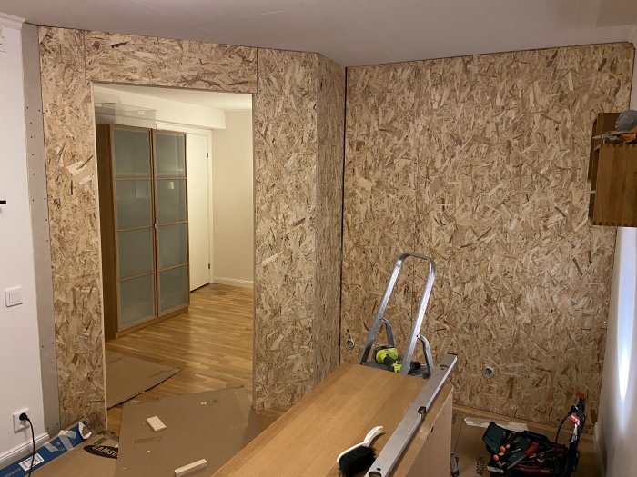 Hörn av ett rum under renovering med OSB-skivor på väggarna, en stege och verktyg i förgrunden.