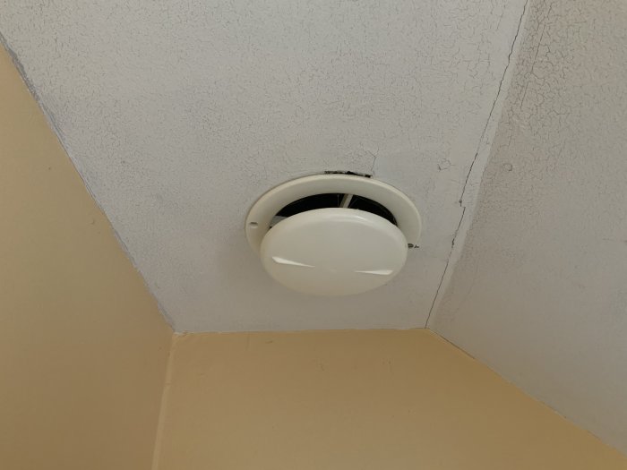 Gammal badrumstakfläkt med sprickor och avflagning på vita taket ovanför gula kakelväggar.