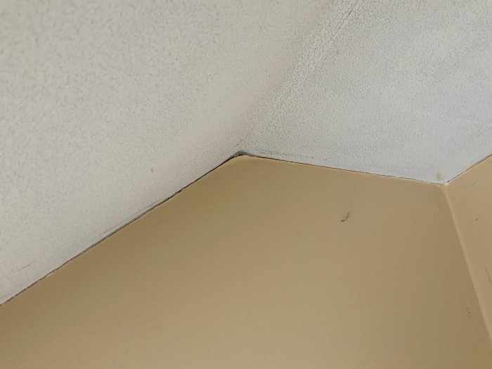 Hörn av gammalt badrum med beige vägg och slitet vitt tak som behöver renovering.