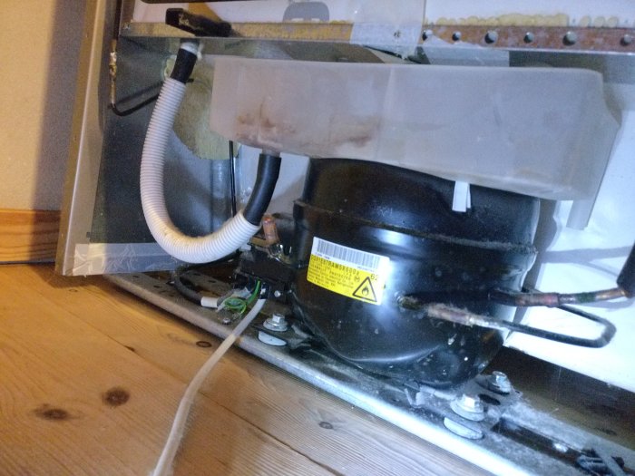 Kompressorn på en Siemens kylfrys med en klämd brödpåseflärp mellan kondenstråget och kompressorn för ljudreducering.