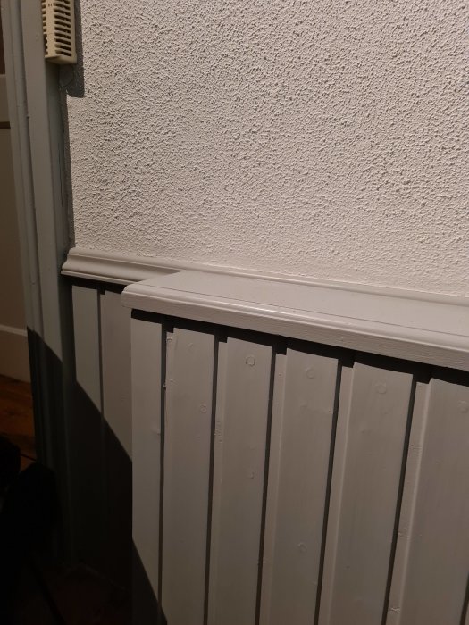 Bild på en hörnsektion av ett rum med vit texturerad vägg och en radiator, samt en termostat ovanför.