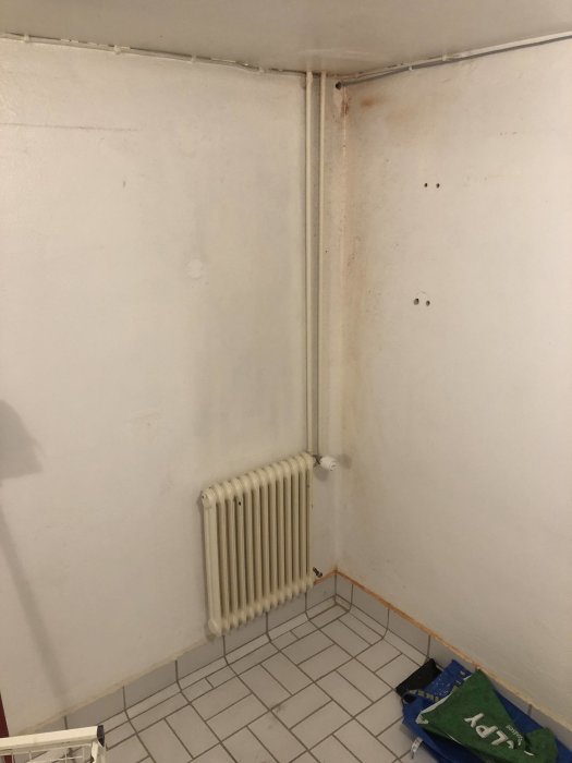 Hörn av en tom tvättstuga med ett radiator-element under ett fönster och rör som går upp längs väggen.