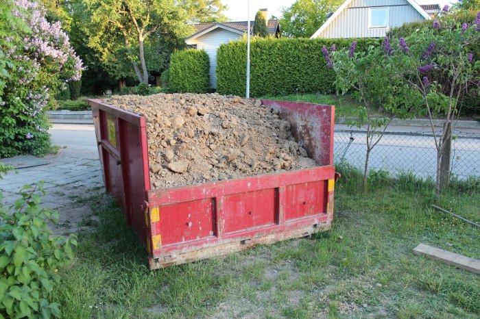 En röd container överfylld med lera placerad på en gräsmatta, med grönska och ett hus i bakgrunden.