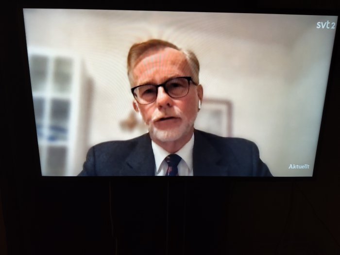 Man i kostym med glasögon intervjuas på tv-skärm, texten "Aktuellt" synlig.