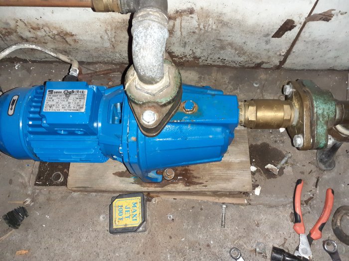 Blå vattenpump monterad i källare, med rörkopplingar och verktyg på golvet, tecken på underhållsarbete.