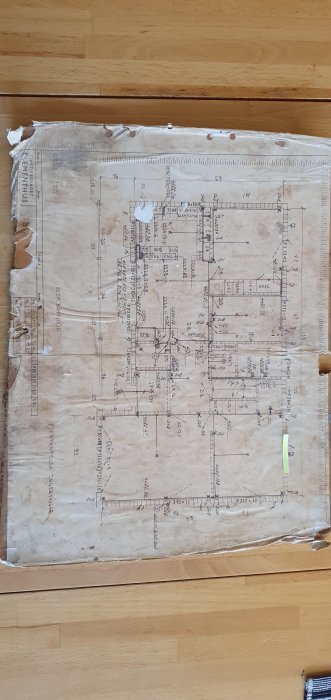 Sliten ritning av en husplan på ett Mockfjärds Elementhus från 1965 placerad på ett trägolv.