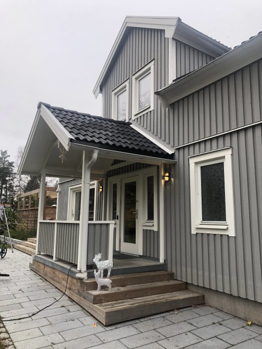 Farstukvist på ett grått hus med betongplattor, trätrappsteg, granitgång och en vit dekorativ ren.