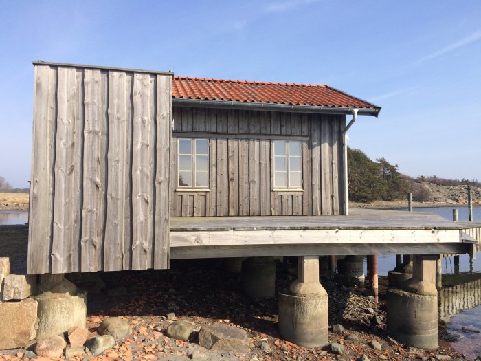 Grå trästuga med röd tegeltak byggd på pålar över strandkanten vid vatten.