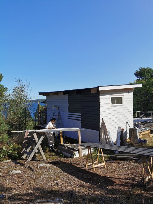 Person målar en bastustuga i matchande färg med garaget, omgivet av byggmaterial och natur.