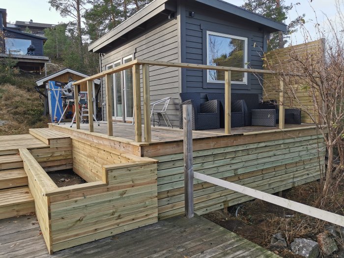 Trädäck vid gråmålat hus med stor blomlåda, räcke under konstruktion och terrassbelysning.