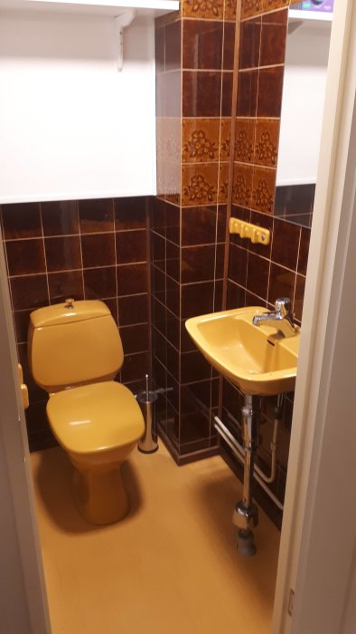 Toalettrum i 70-talsstil med brunt kakel, en beige toalett och handfat, samt en delvis vit övermålad vägg.