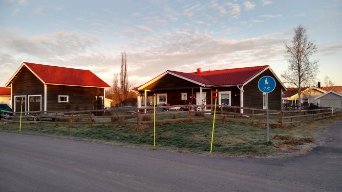 Röd och brun trähus med röda tak i morgonsol vid en gång- och cykelvägsskylt. Frost på gräset.