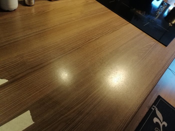 Skador och jack på brun köksbänk i träimitation bredvid svart spishäll, fråga om reparation.