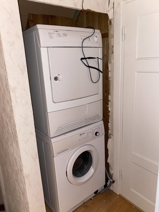 Staplade vitvaror, en torktumlare ovanpå en tvättmaskin, inskjutna i en råväggsgarderob, bredvid en vit dörr.