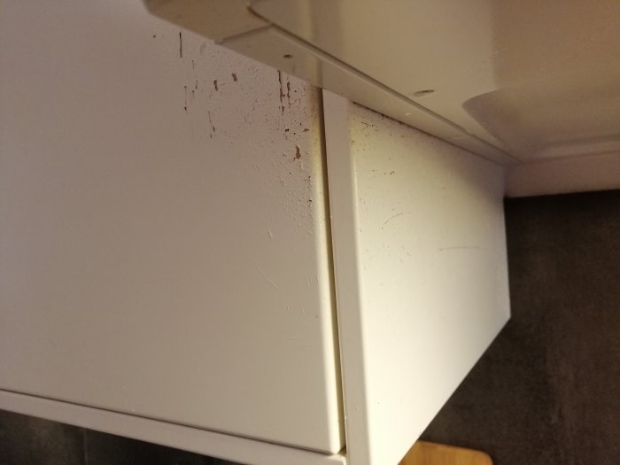 Skador på vita köksluckor med flagnad färg nära ventilationsfläkten och ugnen.