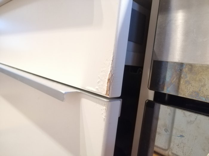 Skador på vita köksluckor nära fläkt och ugn, ytbeläggningen har flagnat och spruckit.