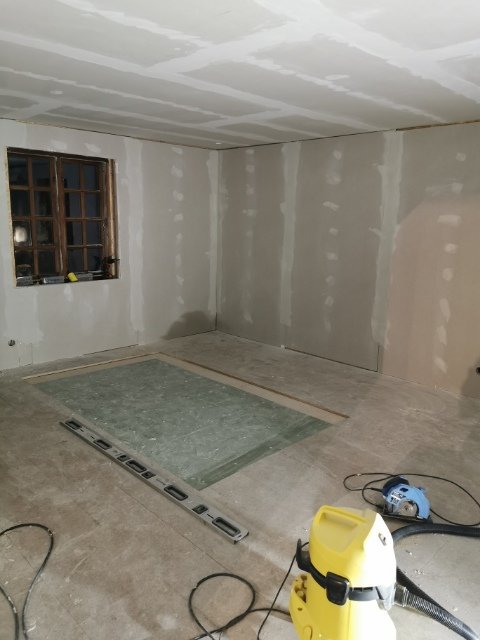 Rum under renovering med spacklade väggar och tak, verktyg på golvet, och ett fönster.