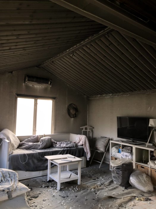 Skador från brand i vardagsrum med sotiga väggar och tak, smält elektronik och rester på golvet.