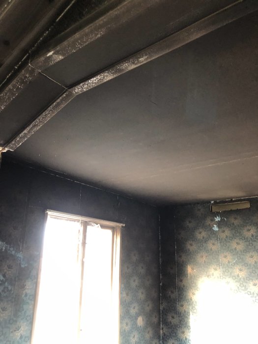 Brandskadat rum med sotiga väggar och innertak, smält plastdetaljer nära fönster i fritidshus från 60-talet.