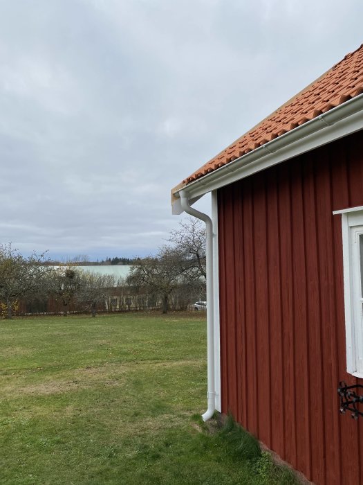 Hörn av ett rött trähus med tegeltak och vit stupränna mot en bakgrund av en grön gräsmatta och träd.