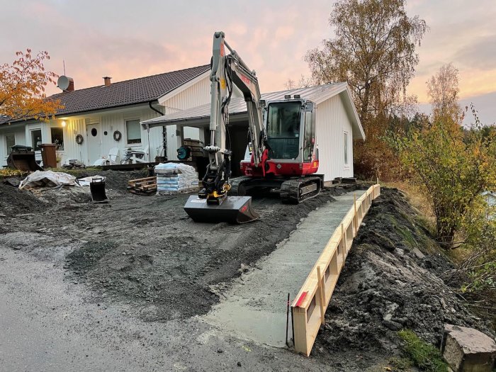 Nygjuten grund för husutbygge med en grävmaskin och byggmaterial i trädgården under skymningen