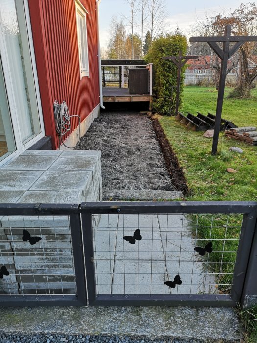 Grusgång anlagd längs rödmålade huset mot en nybyggd träaltan, med ett staket inramat av fjärilsmönster i förgrunden.