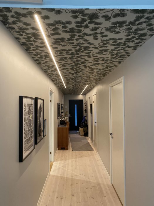Lång hall med LED-stripes på taket, mönstrad tapet och vitmålade väggar, inramad av flera dörrar och tavlor.