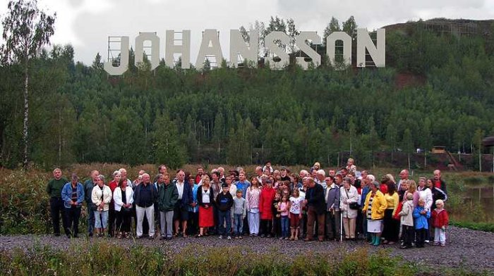 Stor grupp människor poserar för ett gruppfoto utomhus med efternamnet "Johansson" i bakgrunden.