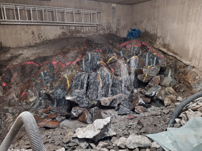 Berggrund med märkta spricklinjer och stenblock i en utgrävd del under en byggnad, med en metallräcke ovanför.