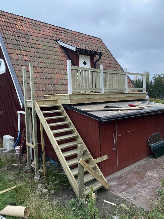 Nybyggd trätrappa utomhus som leder upp till en altan på ett rött hus med tegeltak.