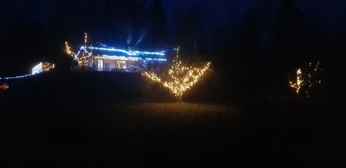 Hus med blå lysande julbelysning och ljusdekorerade träd i skymning, rök från skorstenen.