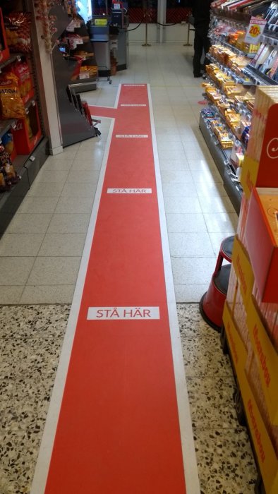 Röd markering på butiksgolv med texten "STÅ HÄR" för fysisk distansering i köområde.