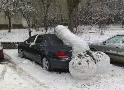 Bil med enorma snöbollar på taket som liknar snögubbar, i ett vinterlandskap.