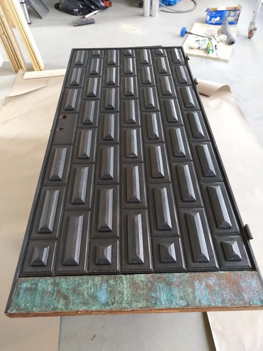 Ny målad svart ytterdörr i teak med dekorativ relief, lutande på golvet i ett byggområde.