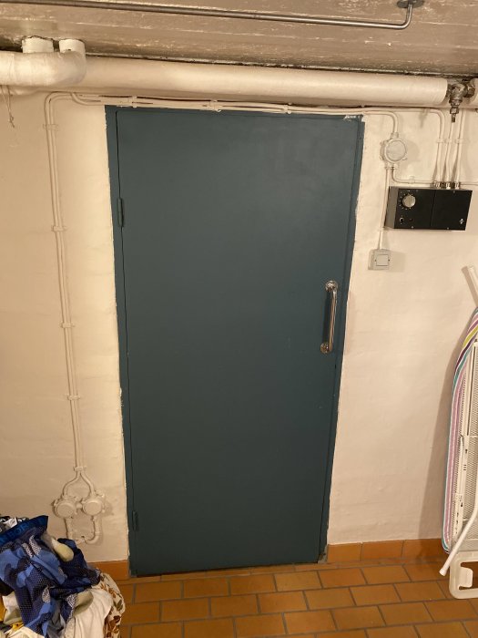 Bastudörr i källare med gammal styrpanel på väggen, vita rör längs väggen, och tegelgolv.