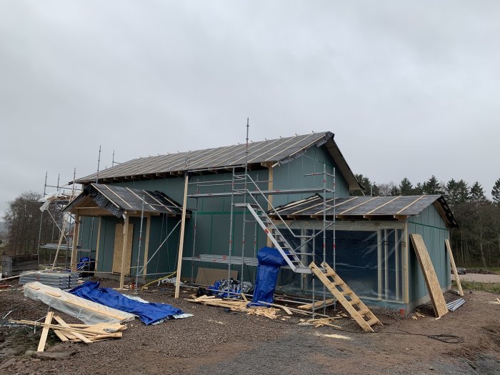 Nybyggt hus i dagsljus med pågående arbete på entrétak, byggställningar och material runtomkring.